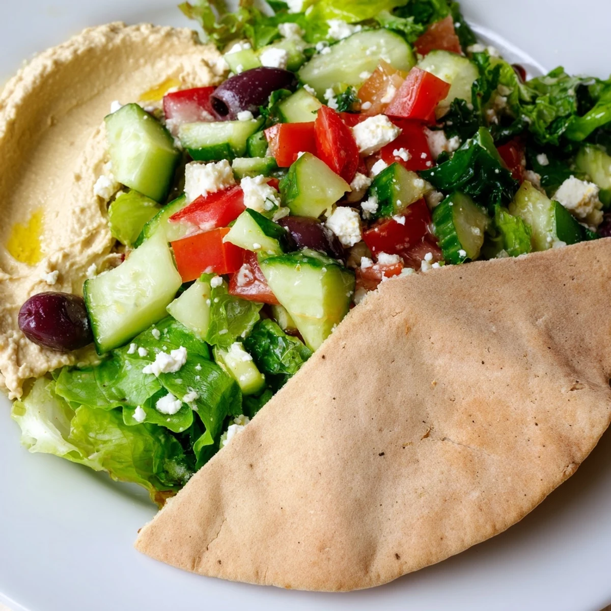 Colorful Mediterranean Salad with Hummus and Pita arranged in a white bowl, topped with feta and olives, with hummus and pita on the side. 