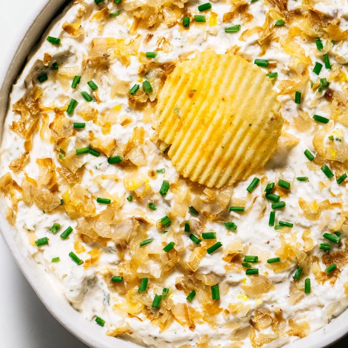 Creamy, golden caramelized onion dip in a bowl, garnished with fresh chives and served next to a pile of crispy potato chips for game day.