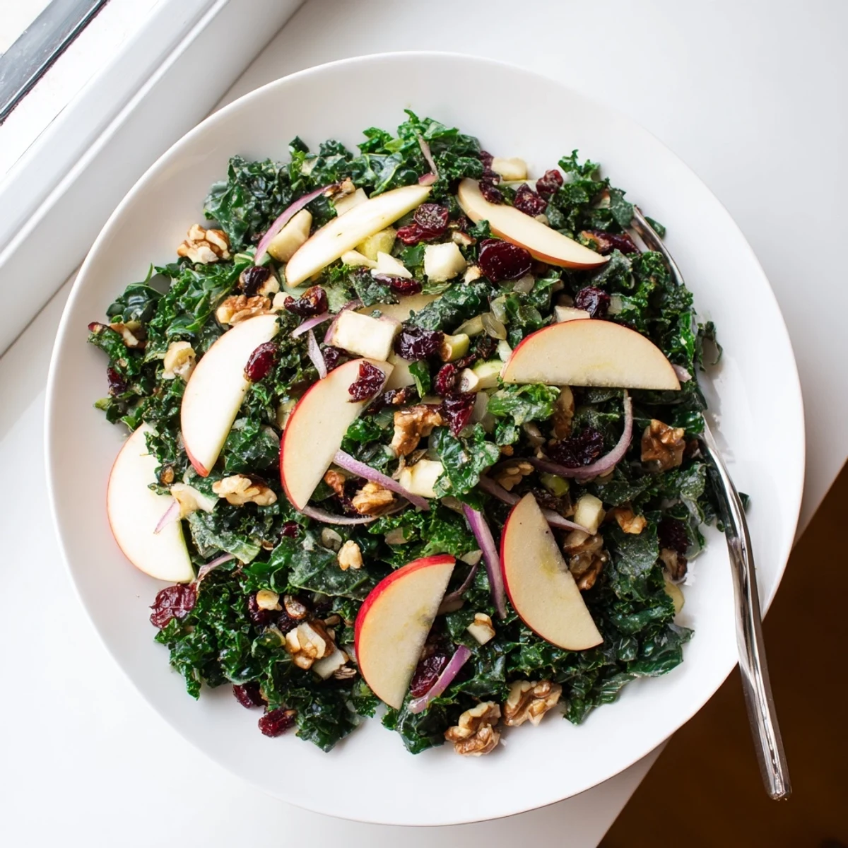 Winter Kale Salad with Apple and Cheddar rests on a white plate, featuring massaged kale leaves, bright apple pieces, red onion slivers, and crunchy nuts for a hearty winter meal.