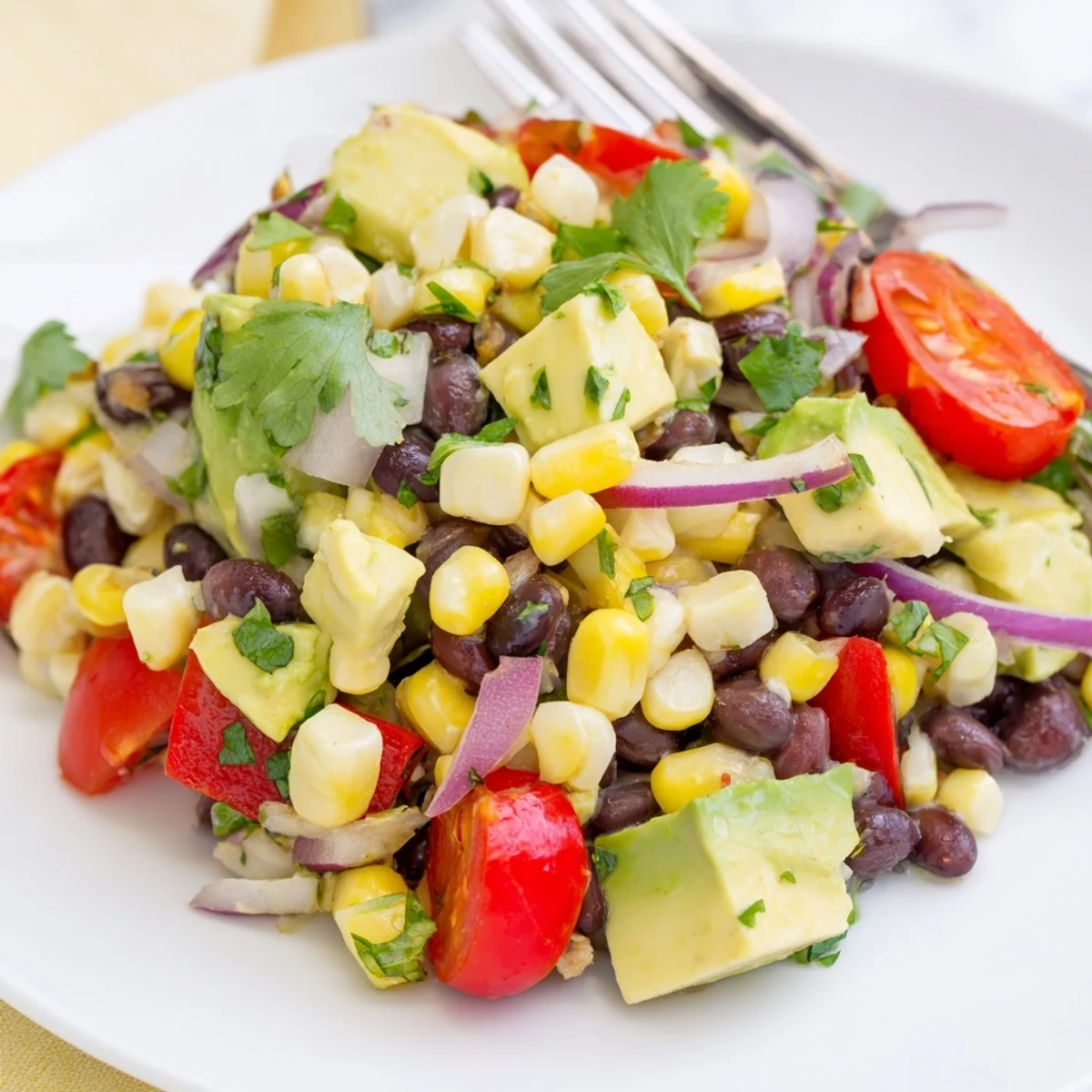 Fresh corn and black bean salad in a white bowl with vibrant red bell peppers, cherry tomatoes, and green cilantro garnish on a wooden table.