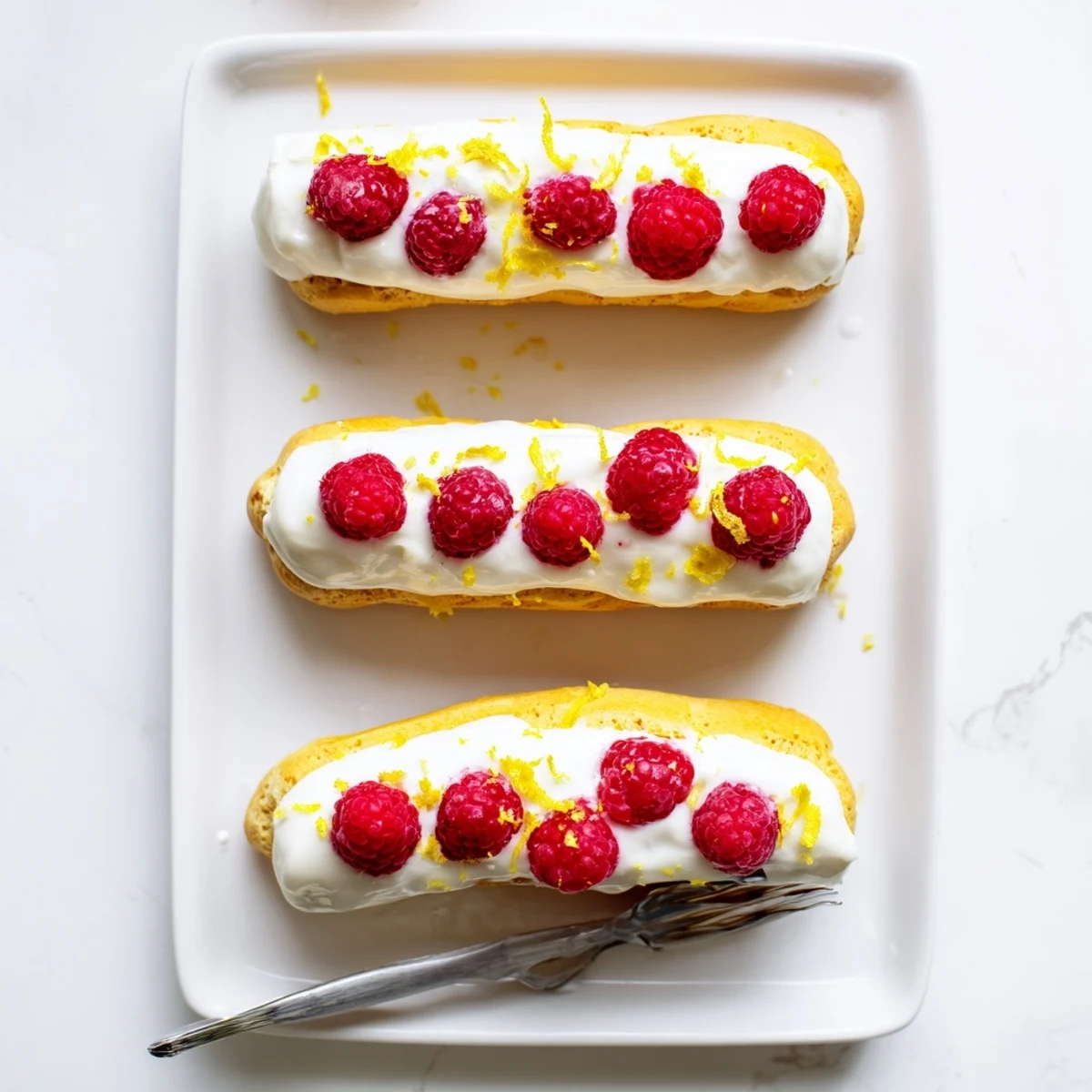 Golden lemon raspberry éclairs filled with tangy cream and topped with fresh raspberries on a white plate