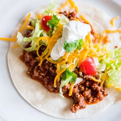 A platter of assembled Beef Taco Night with fresh lettuce, diced tomatoes, shredded cheddar, and a dollop of sour cream.